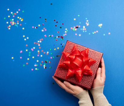 female hands holds a beautiful shiny red box with a big red bow on a blue background with multi-colored sparkles, concept of congratulations and surprise, top view