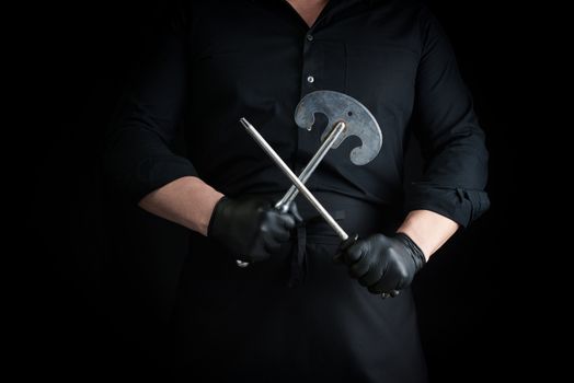 male cook in black uniform and black latex gloves holds a large sharp vintage knife for meat and vegetables and a metal sharpener for knives, black background