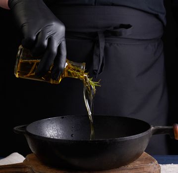 chef in black latex gloves pours olive oil from a transparent bottle into a black cast-iron frying pan, close up
