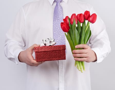 adult man in a white shirt and a lilac tie holding a bouquet of red tulips with green leaves and gift box on a white background, concept for happy birthday, anniversary, valentines day