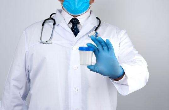 male doctor in a white coat and tie stands and holds a empty plastic container for urine specimen, wearing blue sterile medical gloves