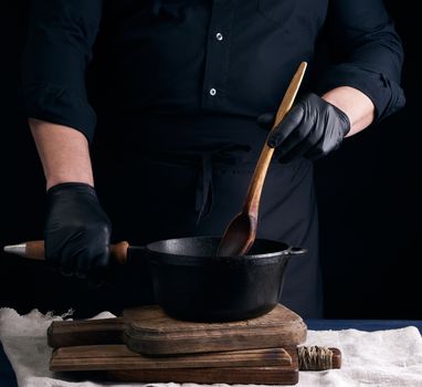 male cook in black uniform and latex gloves stirs a vintage wooden spoon food in a cast-iron pan with a handle, dark background
