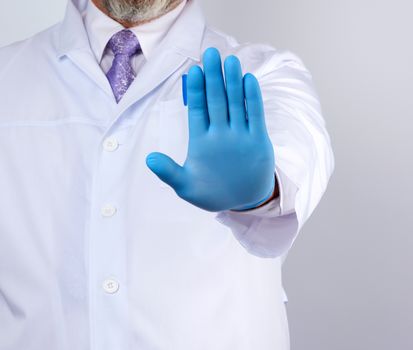 male doctor in a white coat and blue sterile gloves shows a stop gesture with his right hand, white background