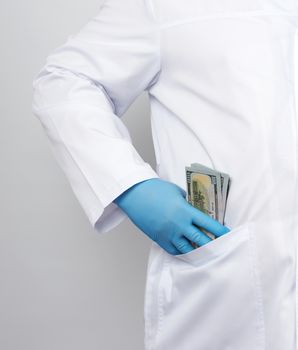 male doctor in a white medical coat and blue latex gloves puts a stack of US dollars in his coat pocket, concept of a bribe