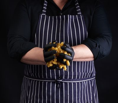 male cook in a striped apron and black latex gloves hold raw fusili pasta, dark background 