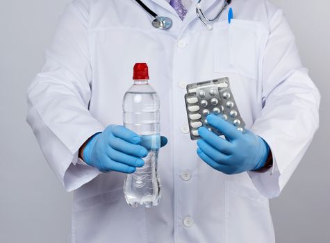 adult doctor therapist is dressed in a white uniform coat and blue sterile gloves is standing and holding a stack of pills in blister packs, white background