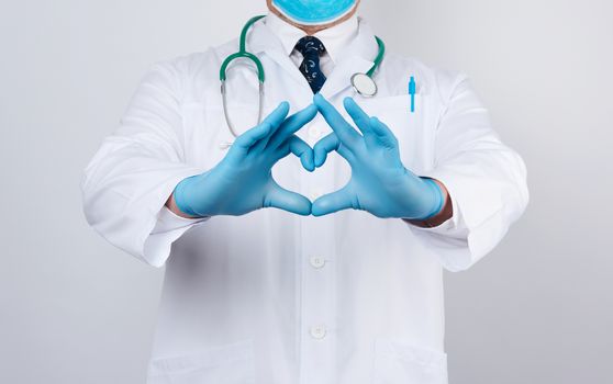 adult doctor man in a white medical coat with a stethoscope on his neck shows a heart gesture with his hands, wearing blue gloves on his hands, white background