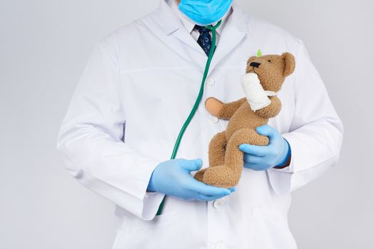 pediatrician in a white coat, blue latex gloves holds a brown teddy bear with a bandaged paw, toy treatment