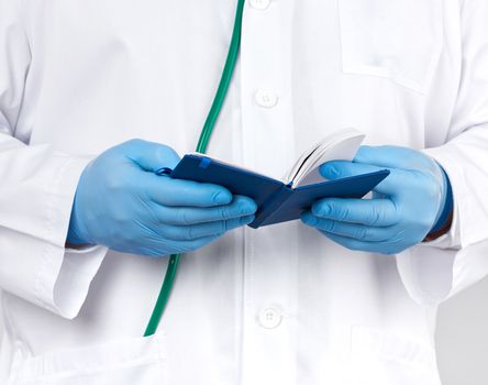 doctor in a white coat and blue gloves holds a blue paper notebook for notes, stethoscope hangs on his neck, white medical backdrop