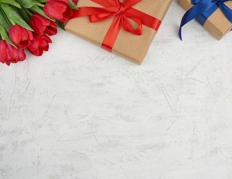 bouquet of red blooming tulips with green leaves, wrapped gifts in brown craft paper and tied with silk ribbon on a white background. Festive backdrop for birthday, Valentine's day, anniversary