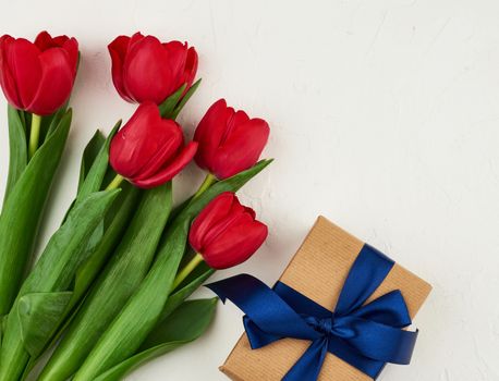 bouquet of red blooming tulips with green leaves, wrapped gift in brown craft paper on a white background, copy space. Festive backdrop for birthday, Valentine's day