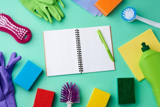 open notebook, rubber gloves for cleaning, multi-colored sponges, brushes and cleaning fluid in a green plastic bottle on a green background, place for text, flat lay