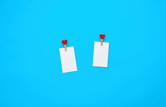 white empty paper rectangular business cards on decorative clothespins with a red heart lie on a blue background, place for an inscription