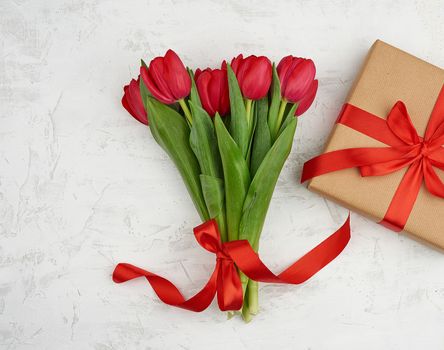 bouquet of red blooming tulips with green leaves, wrapped gift in brown craft paper and tied with a silk red ribbon on a white background, top view. Festive backdrop for birthday, Valentine's day, anniversary