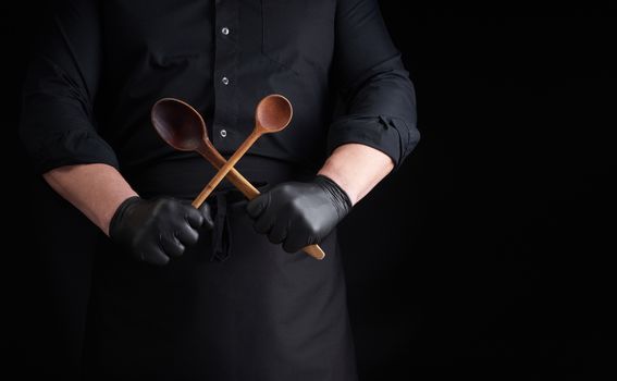 chef in black latex gloves and a black uniform holds crossed wooden vintage spoons in front of him, low key, copy space