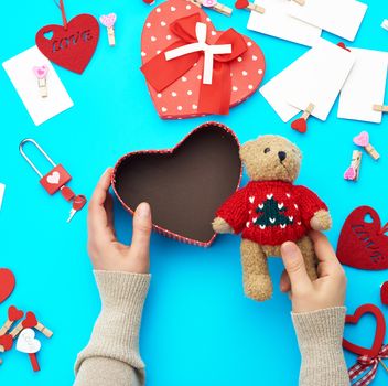 female hands holding a small brown teddy bear, on a blue background an open empty heart-shaped box, top view, holiday Valentine's Day