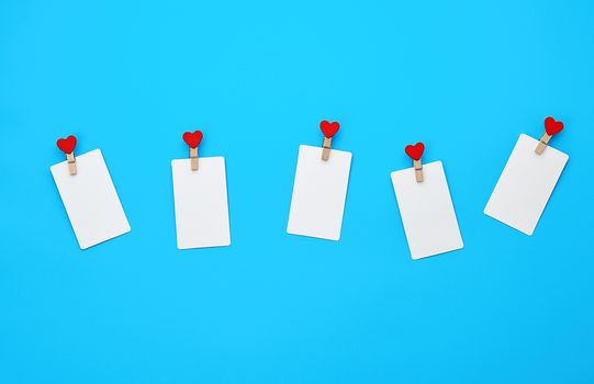 white empty paper rectangular business cards on decorative clothespins with a red heart lie on a blue background, place for an inscription