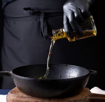 chef in black latex gloves pours olive oil from a transparent bottle into a black cast-iron frying pan, close up