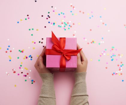 two female hands in a sweater hold closed square pink box with a bow on a pink background, top view, festive backdrop