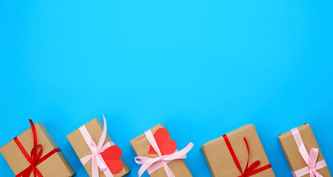 box is wrapped in brown paper and tied with a red thin silk ribbon on a blue background, red cut out paper hearts, copy space, holiday is Valentine's Day