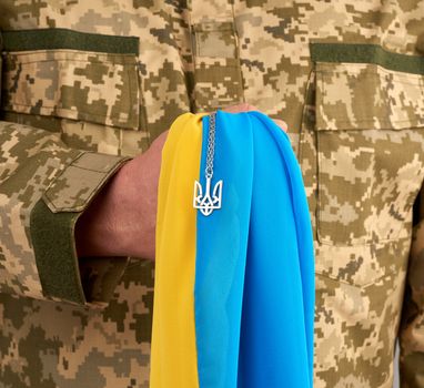 Ukrainian man warrior dressed in a military pixel uniform holds the yellow-blue flag of the state of Ukraine and on the chain a small coat of arms of the country of trident, close up