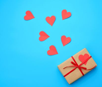 box is wrapped in brown paper and tied with a red thin silk ribbon on a blue background, red cut out paper hearts, copy space, holiday is Valentine's Day