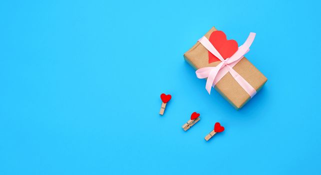 box is wrapped in brown paper and tied with a red thin silk ribbon on a blue background, red cut out paper hearts, copy space, holiday is Valentine's Day