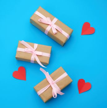 box is wrapped in brown paper and tied with a red thin silk ribbon on a blue background, red cut out paper hearts, copy space, holiday is Valentine's Day