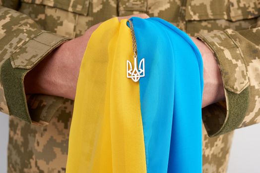 Ukrainian man warrior dressed in a military pixel uniform holds the yellow-blue flag of the state of Ukraine and on the chain a small coat of arms of the country of trident, close up