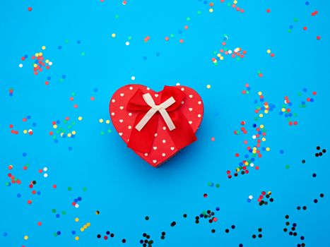 closed red heart-shaped cardboard box on a blue background with multi-colored shiny confetti, festive backdrop for birthday, Valentine's day