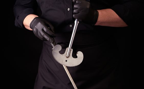 male cook in black uniform and black latex gloves holds a large sharp vintage knife for meat and vegetables and a metal sharpener for knives, black background