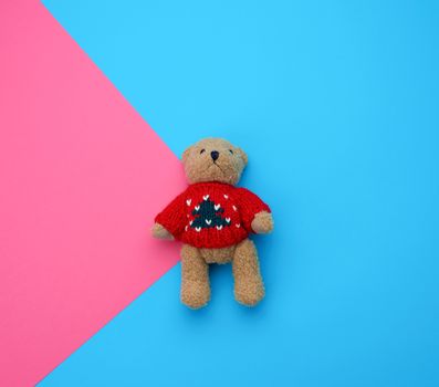 little cute brown teddy bear with in a red knitted sweater on a blue pink background, holiday toy