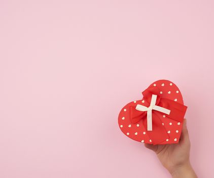 female hand holding a red cardboard box with a bow on a pink background, concept of giving a gift for a holiday