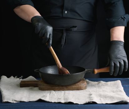 male cook in black uniform and latex gloves stirs a vintage wooden spoon food in a cast-iron pan with a handle, dark background