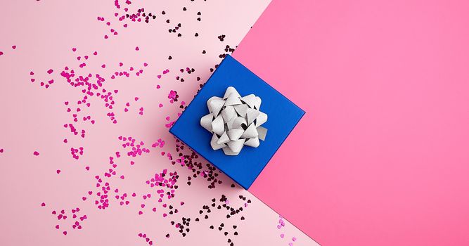 closed square blue gift box with silver bow on a pink background, near multi-colored sequins, top view, festive backdrop
