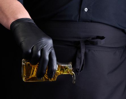 man in black clothes, latex gloves holds a transparent bottle with olive oil, low key