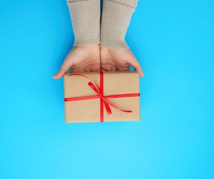 female hand holds a brown box with a red bow on a blue background, concept of a congratulation on a happy birthday, place for text