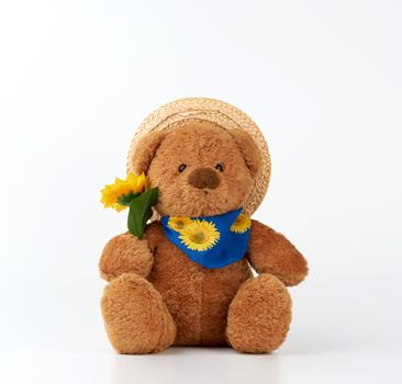 cute little brown teddy bear holds a plastic yellow sunflower and is dressed in a straw hat,  toy is sitting on a white background