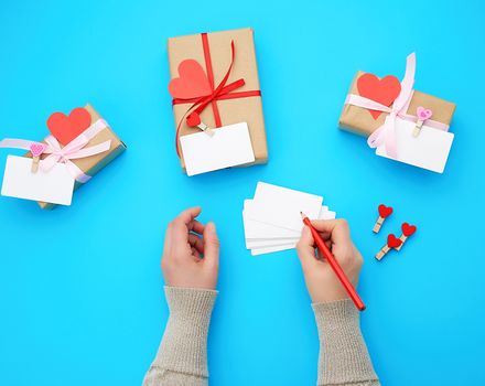 female hand holds a box wrapped in brown kraft paper and tied with a silk red ribbon on a blue background, paper red hearts. Valentine's Day backdrop