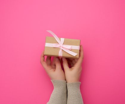 female hand holds a brown box with a red bow on a pink background, concept of a congratulation on a happy birthday, place for text