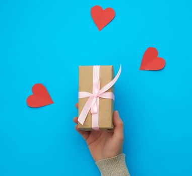 female hand holds a box wrapped in brown kraft paper and tied with a silk ribbon on a blue background, paper red hearts. Valentine's Day backdrop