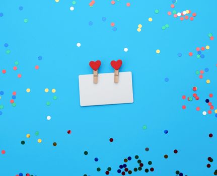 empty white rectangular business card in a wooden clothespin on a blue background with multi-colored contetti, festive backdrop