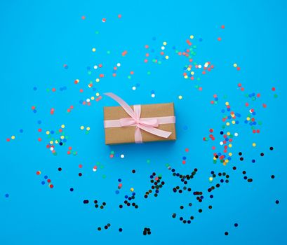 brown gift box wrapped in paper and tied with silk pink ribbon on a blue background with multi-colored shiny confetti, festive backdrop for birthday, valentine's day