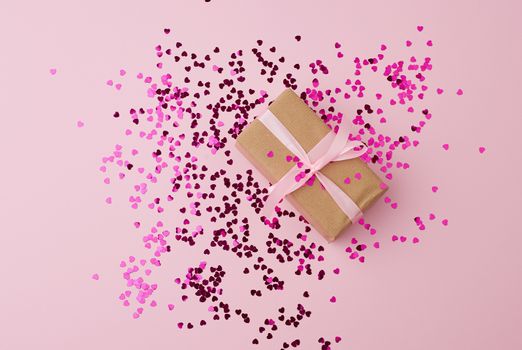 brown gift box wrapped in paper and tied with silk pink ribbon on a pink background with multi-colored shiny confetti, festive backdrop for birthday, valentine's day