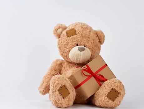 cute big beige teddy bear holds a brown box with a red ribbon on a white background. Festive concept