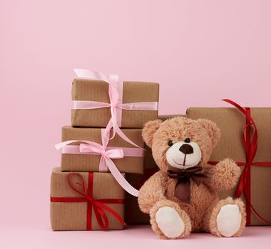 teddy bear and gifts wrapped in brown eco kraft paper and tied with a ribbon on a pink background, festive backdrop for Valentine's day, birthday