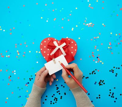female hand holds a red box and tied with a silk red ribbon on a blue background. Valentine's Day backdrop, girl signs a business card