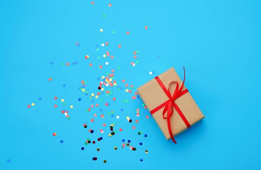 brown gift box wrapped in paper and tied with silk ribbon on a blue background with multi-colored shiny confetti, festive backdrop for birthday, valentine's day