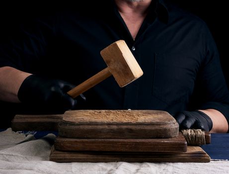 male cook in black uniform holds a wooden hammer for beating meat on a vintage cutting board, black background