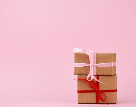 stack of gifts in boxes wrapped in brown kraft paper and tied with silk ribbon on a pink background. Festive concept, copy space